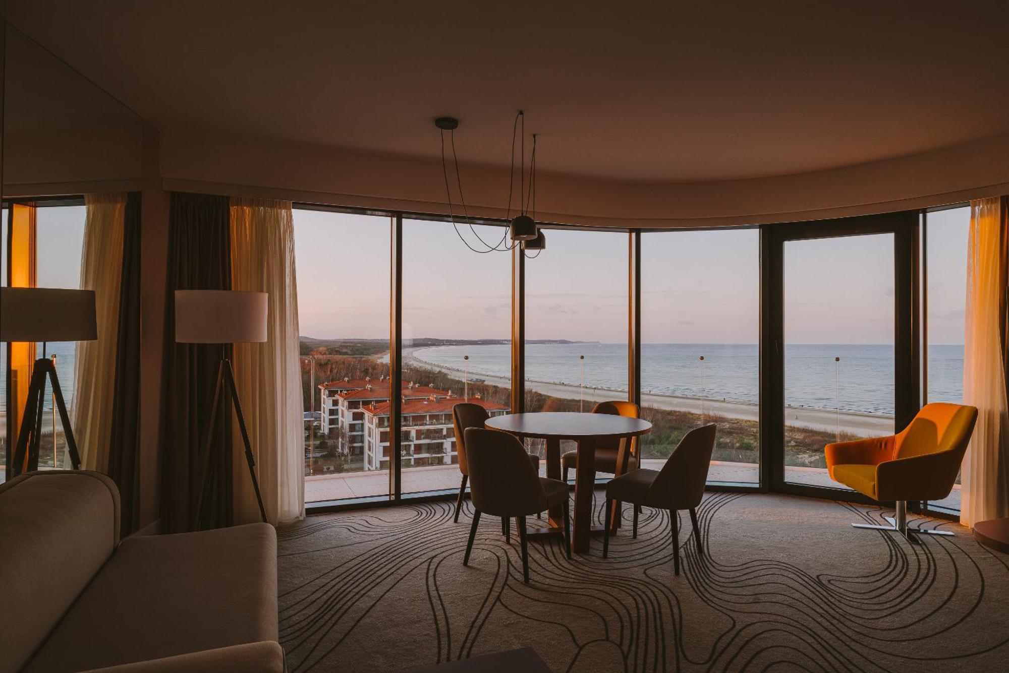 Radisson Blu Resort Świnoujście Zewnętrze zdjęcie The photo shows a modern and stylish interior of a room featuring large windows that provide a view of the beach and ocean. The room is furnished with a round table and chairs, along with a cozy sofa and a bright yellow armchair. The decor has a cont