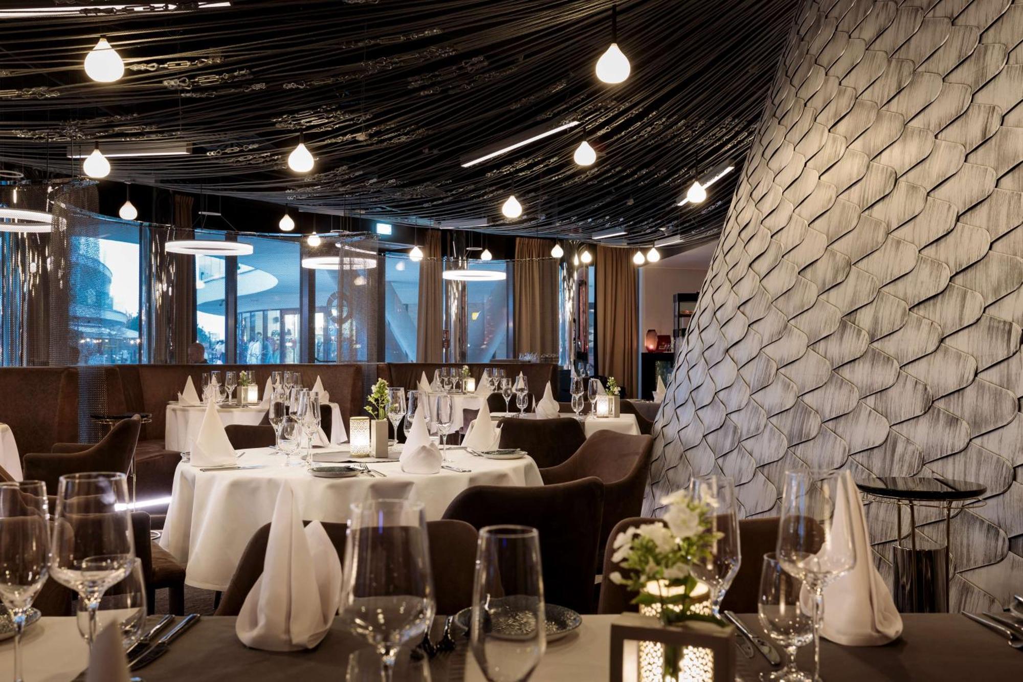 Radisson Blu Resort Świnoujście Zewnętrze zdjęcie The photo shows a stylish restaurant interior. The dining area features elegantly set tables with white tablecloths, glassware, and folded napkins. There are decorative elements like small floral arrangements on the tables. The ceiling is adorned wit