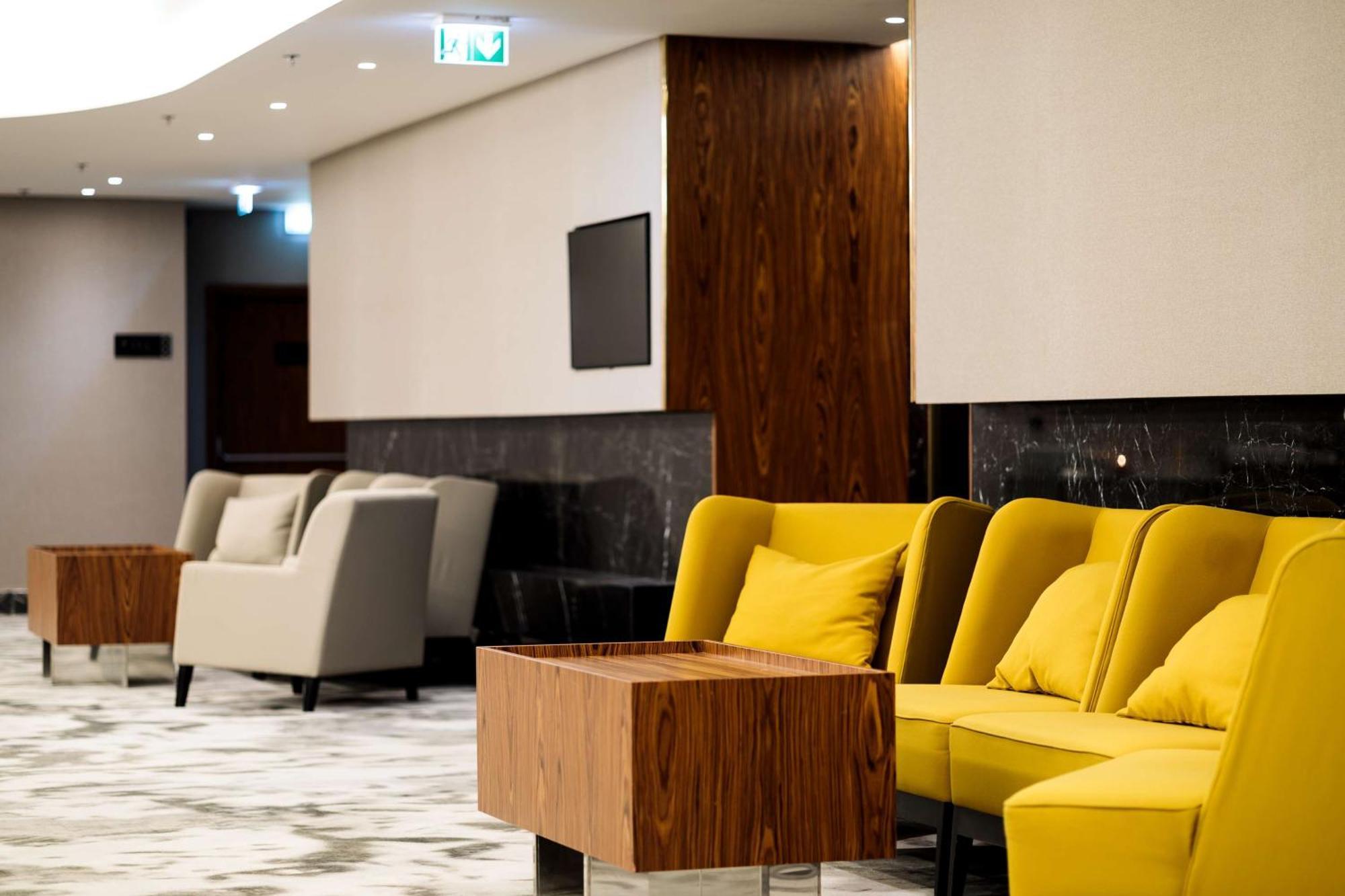 Radisson Blu Resort Świnoujście Zewnętrze zdjęcie The photo depicts a modern lounge or waiting area with stylish seating. On the left, there are soft gray armchairs arranged around a wooden coffee table. In the center, there are yellow chairs with plush cushions, contributing a vibrant contrast to t
