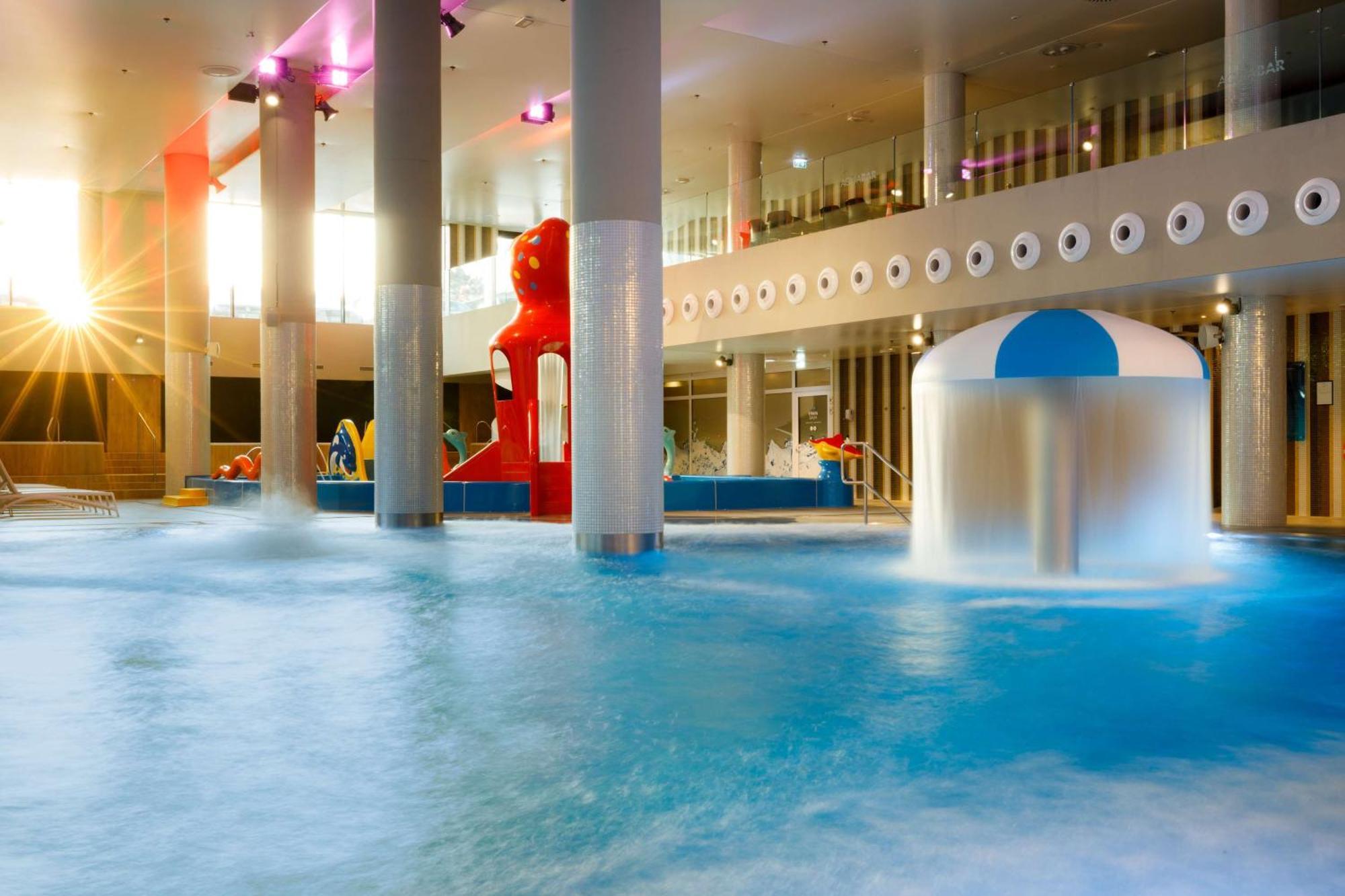 Radisson Blu Resort Świnoujście Zewnętrze zdjęcie The photo shows an indoor water park or pool area. There is a large body of water with playful features, including a water slide shaped like a red bear and a smaller structure with a blue and white canopy. The lighting is bright, suggesting a sunny a