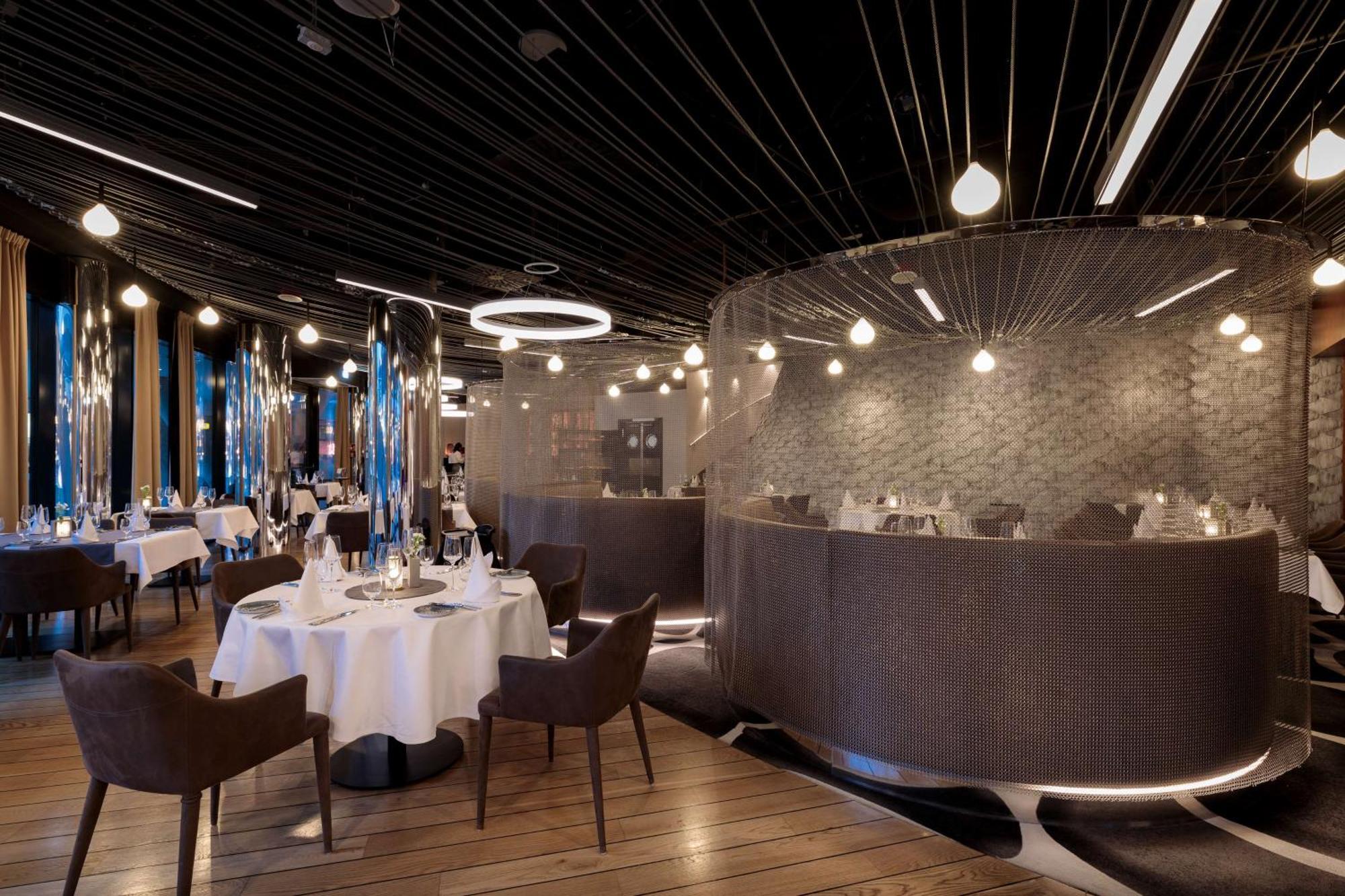 Radisson Blu Resort Świnoujście Zewnętrze zdjęcie The photo shows an elegant restaurant interior. It features polished wooden floors and contemporary decor. Round tables are set with white tablecloths and have chairs arranged around them. The lighting is modern, with multiple pendant lights hanging 