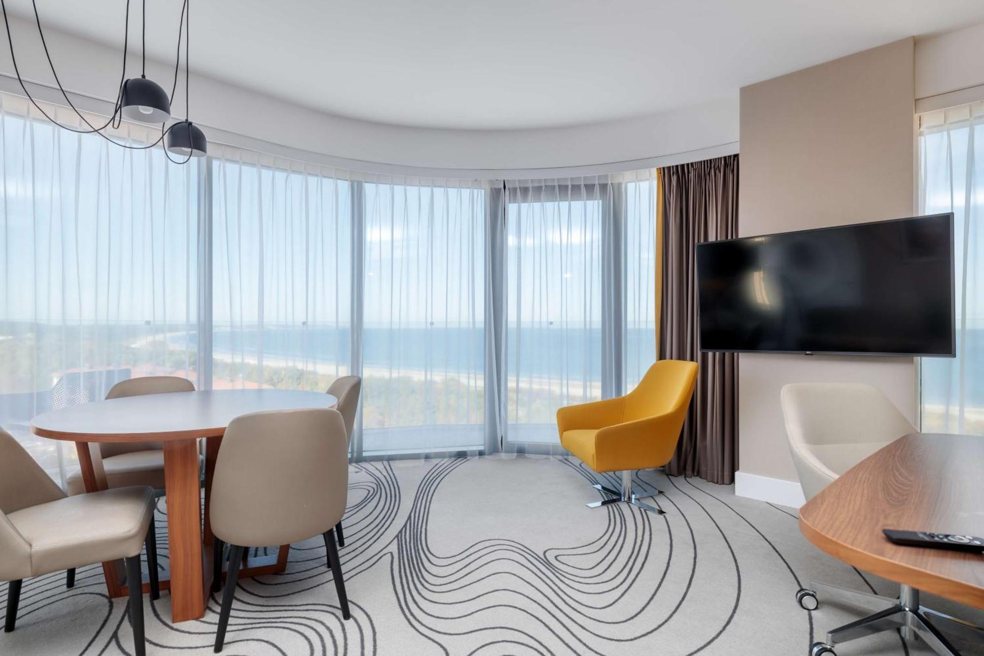 Radisson Blu Resort Świnoujście Zewnętrze zdjęcie The photo shows a modern, stylish interior of a room with large windows that provide a panoramic view. The space features a round dining table surrounded by chairs, including a distinct yellow armchair. There is a flat-screen television mounted on th