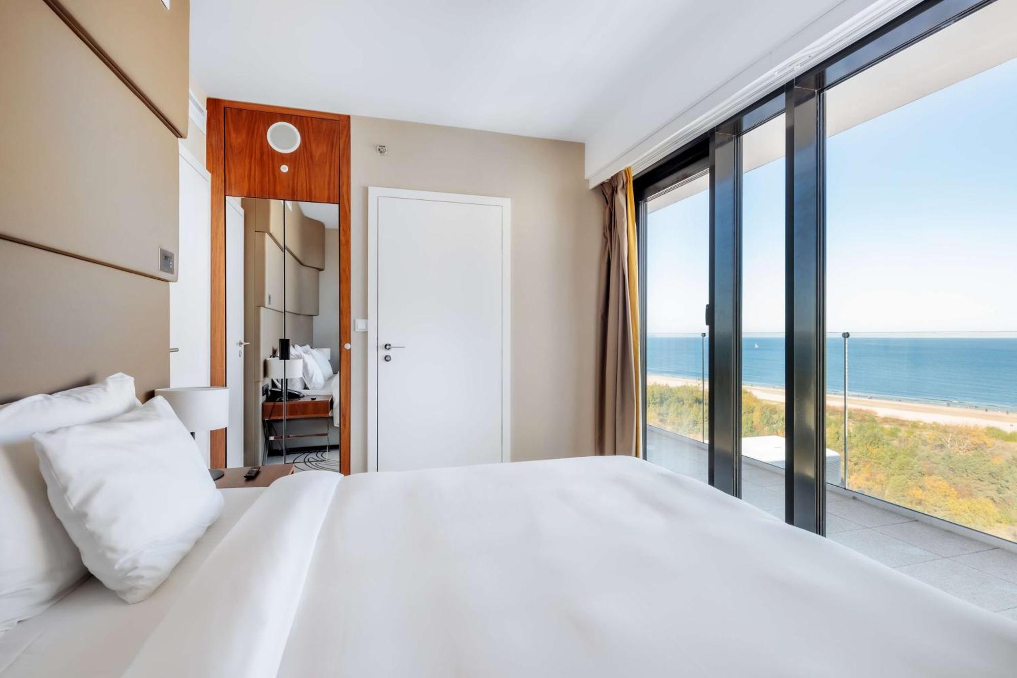 Radisson Blu Resort Świnoujście Zewnętrze zdjęcie The photo shows a modern hotel room with a bright and airy atmosphere. There is a large bed with white bedding positioned near a glass wall that offers a view of the ocean. The room features neutral-colored walls and wooden accents, enhancing its con