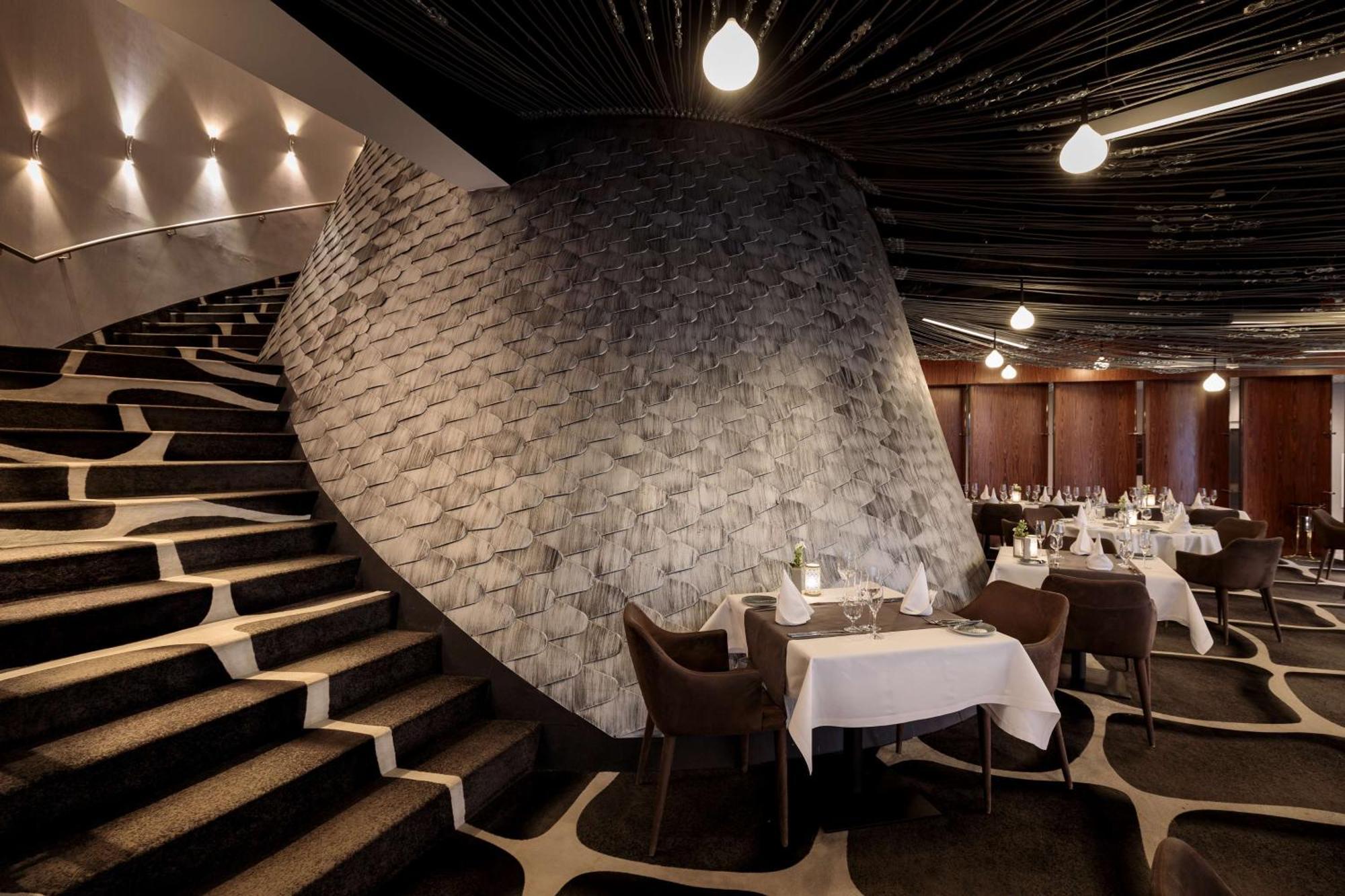 Radisson Blu Resort Świnoujście Zewnętrze zdjęcie The photo shows the interior of a stylish restaurant. In the foreground, there are elegantly set tables with white tablecloths and dark chairs. A unique, curving stone wall adds a distinct architectural feature, and a grand staircase with a textured 
