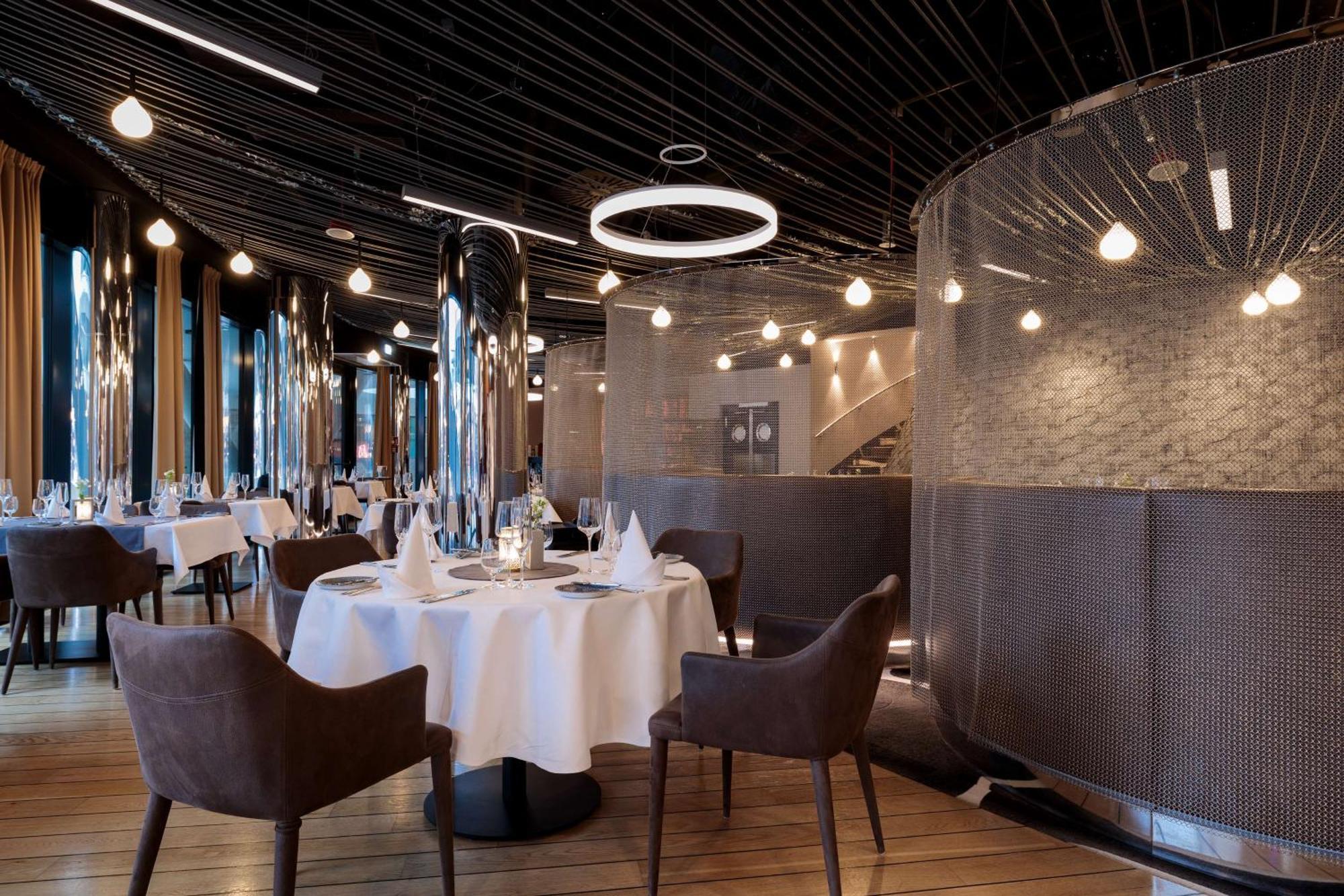 Radisson Blu Resort Świnoujście Zewnętrze zdjęcie The image shows a modern restaurant interior that features a stylish design. There are several round tables with white tablecloths. The seating consists of brown, upholstered chairs. The space is illuminated by various hanging lights, including a dis