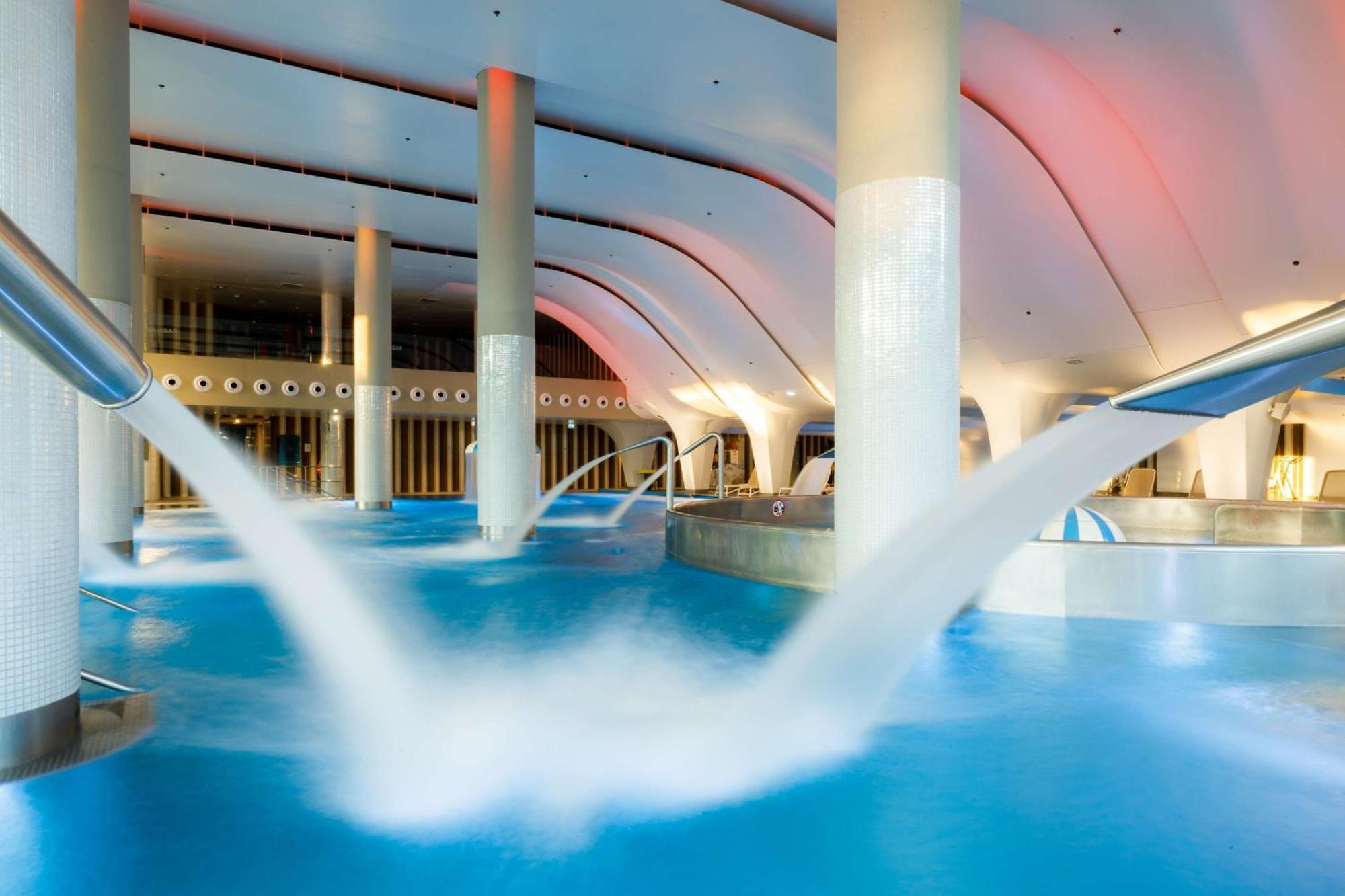 Radisson Blu Resort Świnoujście Zewnętrze zdjęcie The photo depicts an indoor swimming pool area characterized by modern architecture. The pool is filled with clear blue water, and there are several columns supporting the ceiling. Water features, such as jets or fountains, are seen creating flowing 