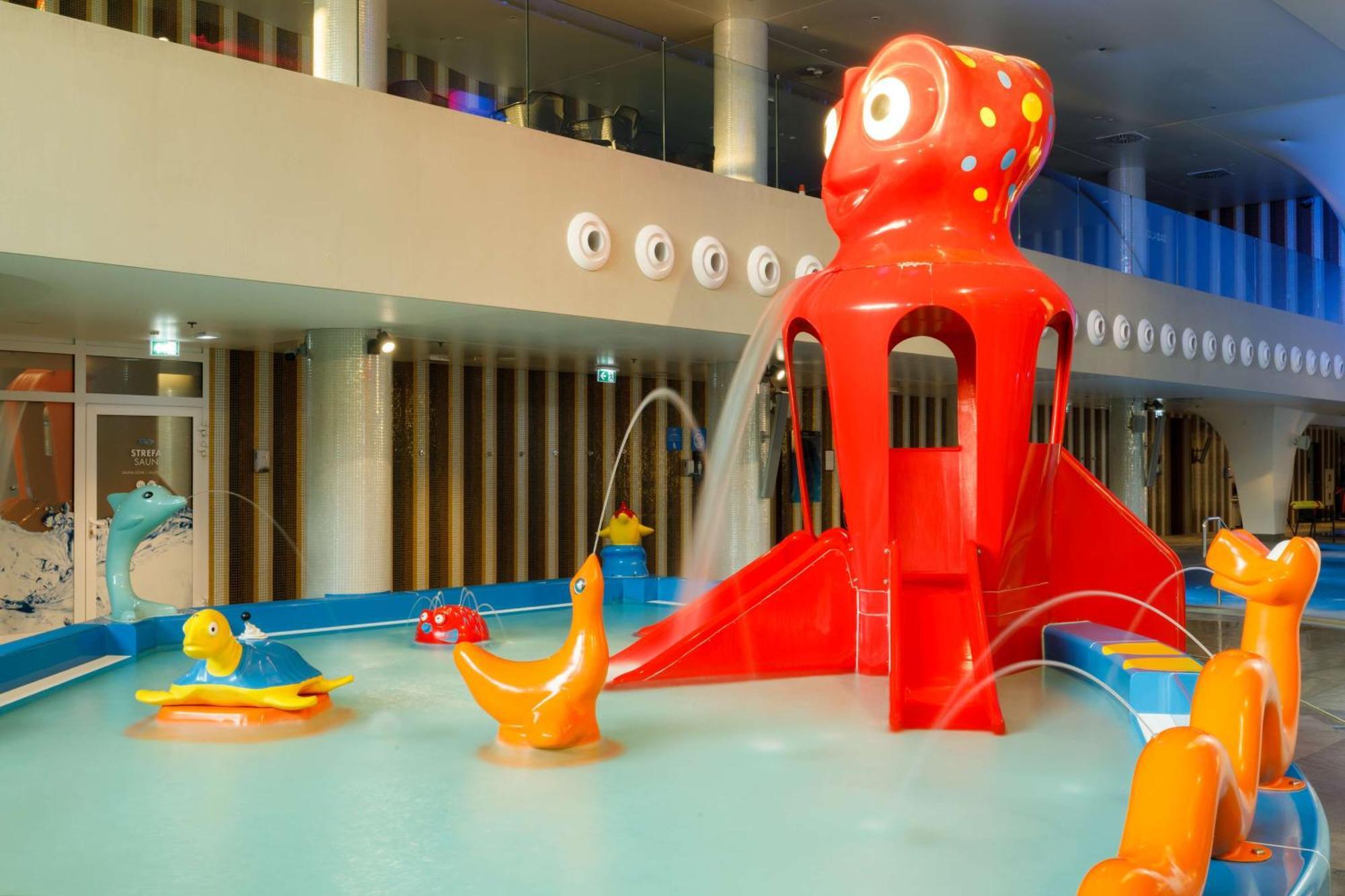Radisson Blu Resort Świnoujście Zewnętrze zdjęcie The photo shows a colorful indoor splash pad or water play area designed for children. Central to the scene is a large, bright red octopus structure, which has water fountains spraying out. Surrounding it are various playful, colorful water animals, 
